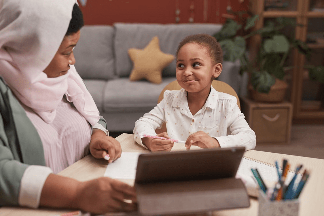 Parent in a hijab helping her daughter with schoolwork on a tablet