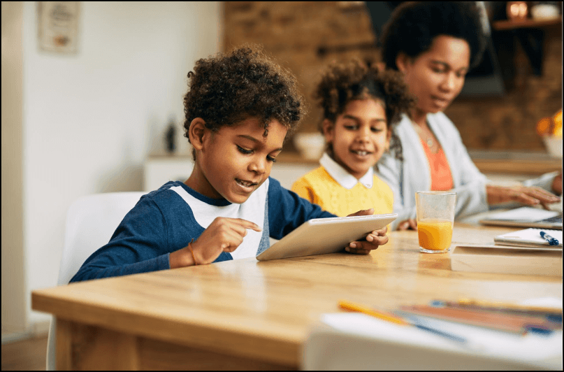 Child pointing at a tablet while a sibling and parent look on at the kitchen table